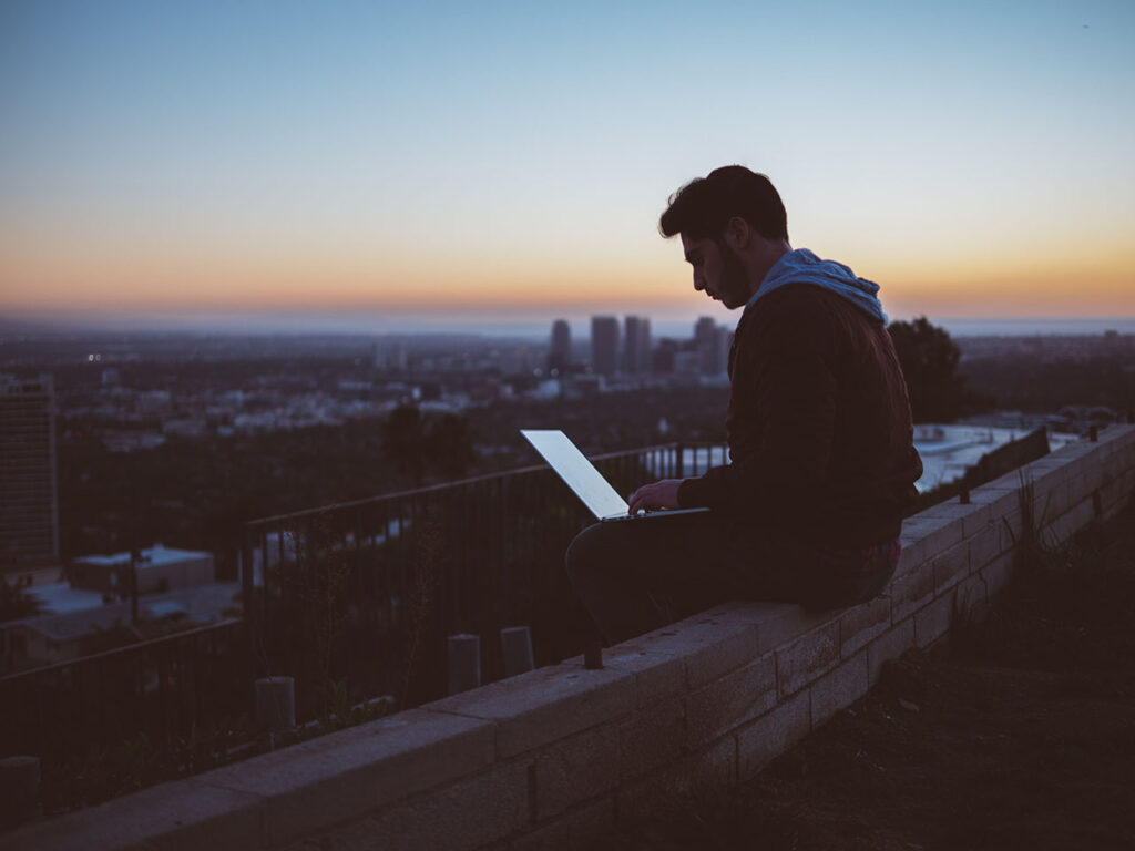 A man sitting on a concrete brick with an open laptop on his lap. © Avi Richards / Unsplash