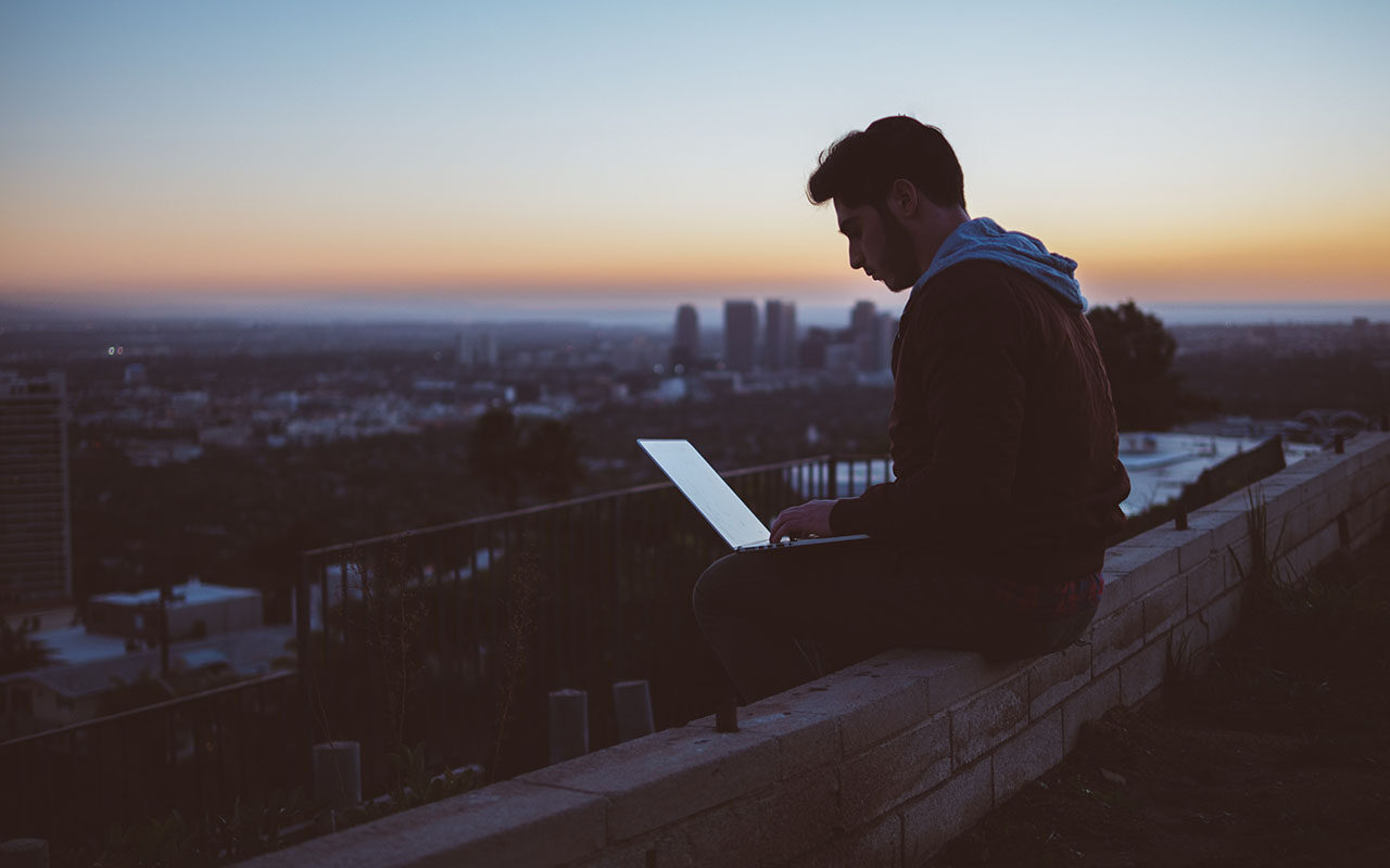 A man sitting on a concrete brick with an open laptop on his lap. © Avi Richards / Unsplash