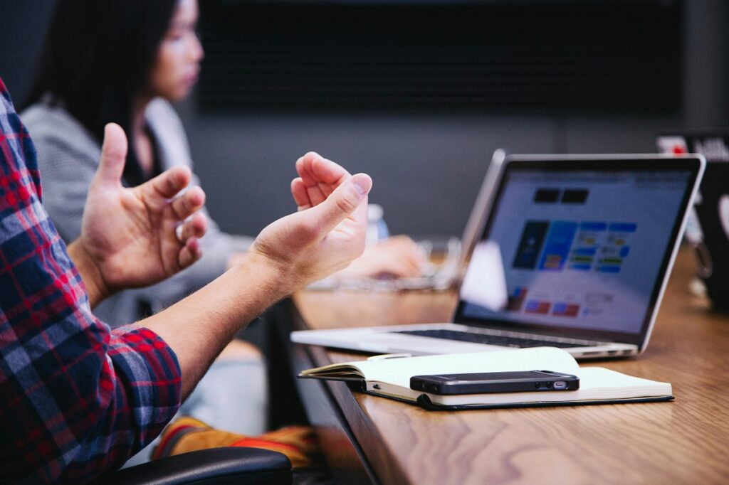 Picture of a man sitting at a table with his laptop open in front of him. His arms are up as if in conversation and you can see a woman colleague in the background.