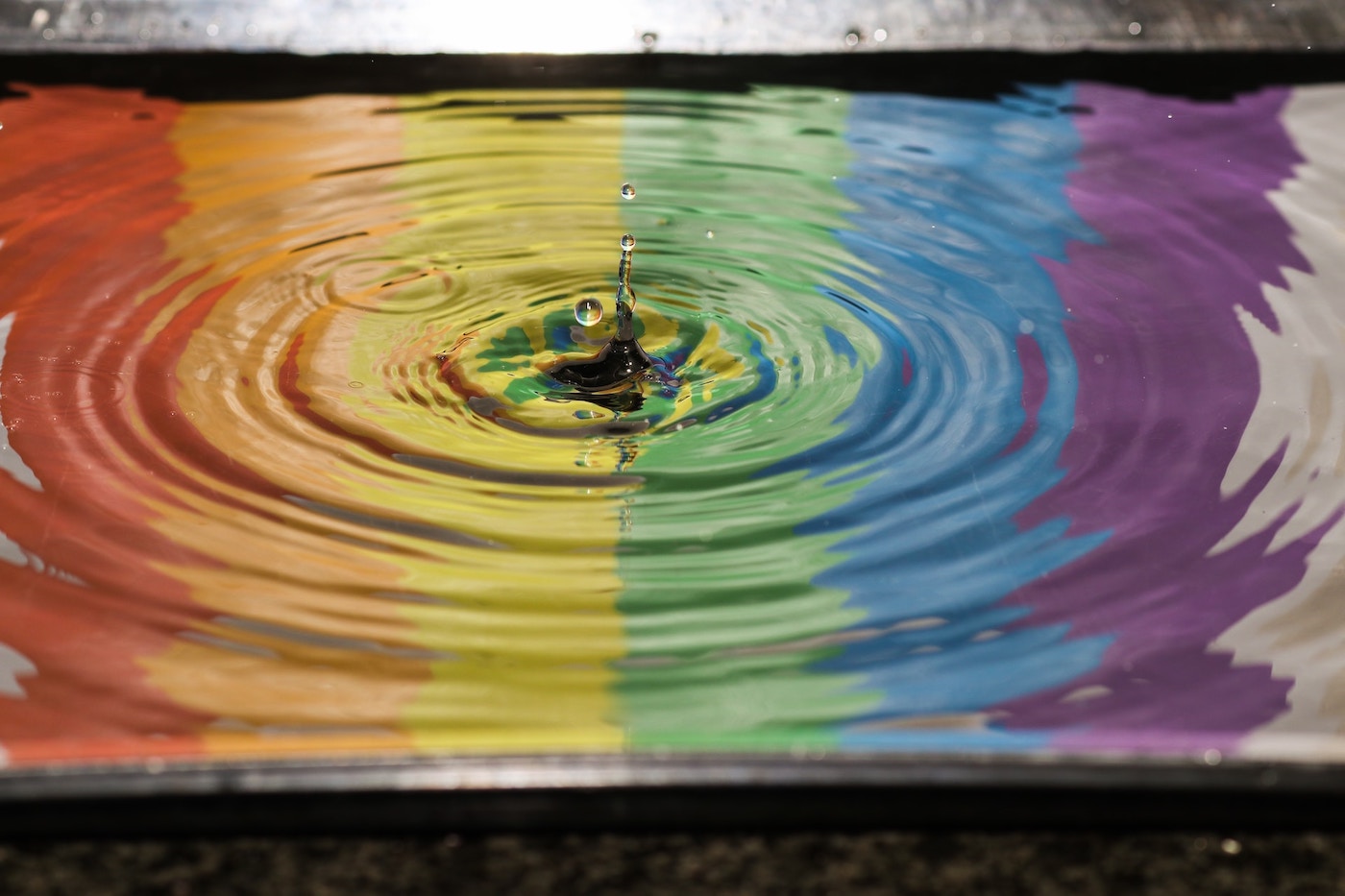 Reflection of a rainbow flag in water with a drop of water causing ripples.