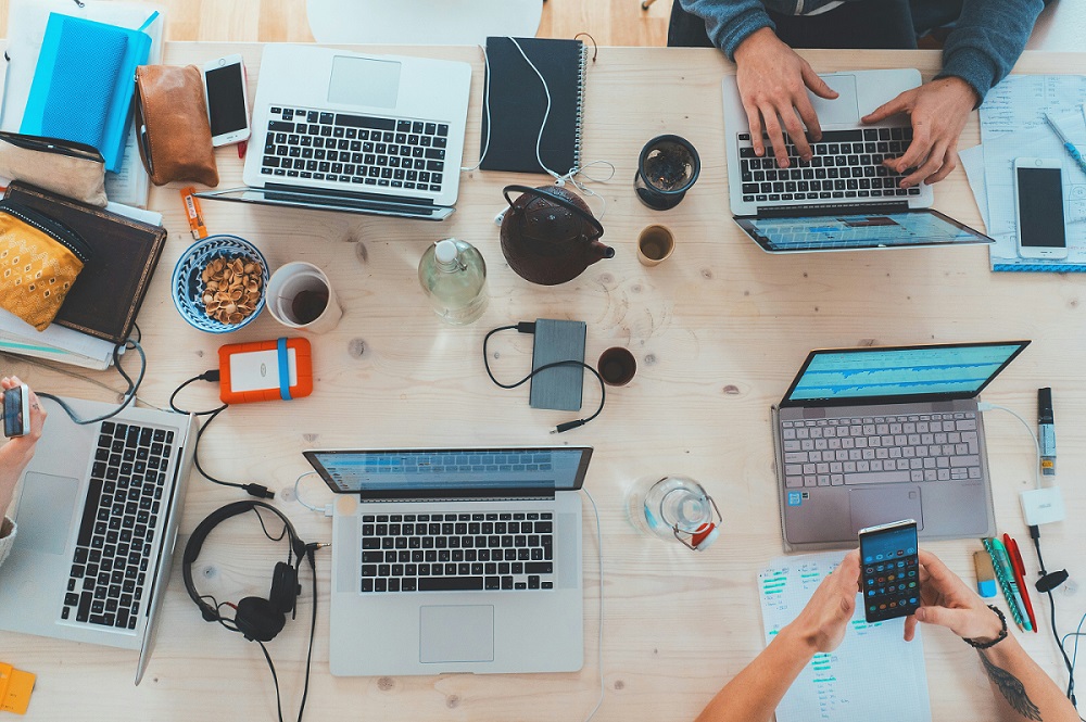 An overhead view of people working together on one big table showing their laptops, snacks, and hands.