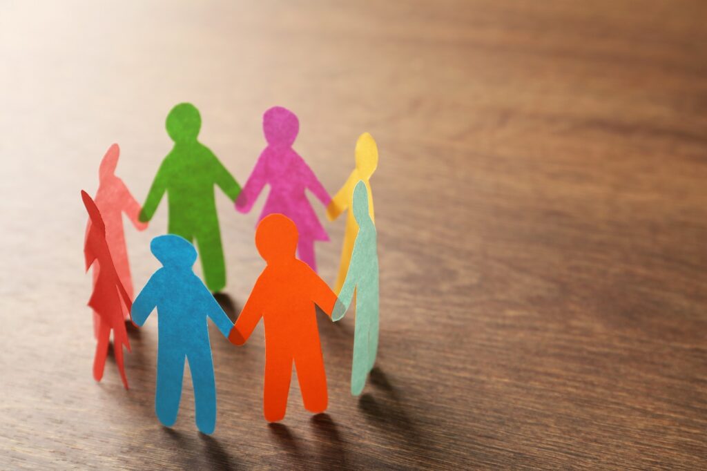 Paper cutouts of people in rainbow colors are stood up in a circle holding hands on a wood surface