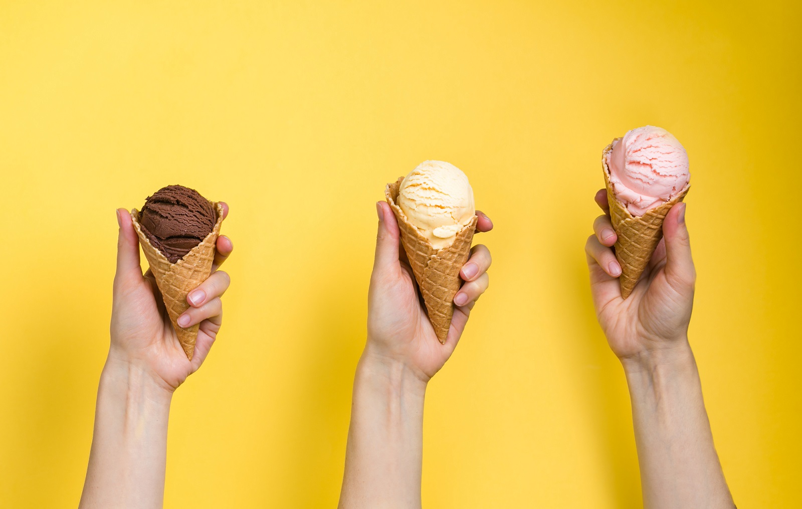 Three hands holding up three different ice cream cones with a scoop of different flavors of ice cream on a yellow background