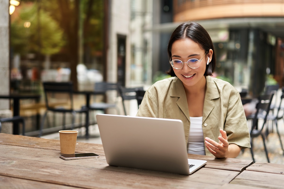 A woman sits in a cafe in front of an open computer, seemingly in the process of doing a video meeting